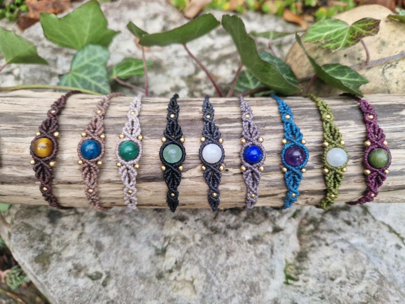 May include: A collection of eight macrame bracelets, each featuring a different gemstone. The bracelets are displayed on a piece of wood, showcasing a variety of colors including brown, blue, green, and purple. The gemstones are round and set in the center of each bracelet.