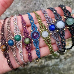 Può includere: Una collezione di braccialetti macramè fatti a mano in vari colori, ognuno con una gemma centrale. I braccialetti sono esposti in una mano, mostrando l'intricato nodo e i diversi colori delle pietre.
