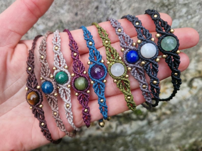 May include: A collection of handmade macrame bracelets in various colors, each featuring a central gemstone. The bracelets are displayed in a hand, showcasing the intricate knotting and the diverse selection of stones, including tiger's eye and amethyst.