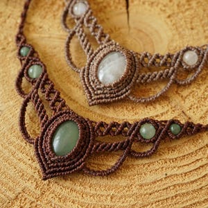 May include: Two handmade macrame necklaces with gemstone accents. One necklace features a large oval green stone, while the other has a large oval white stone. Both are on a wooden surface.