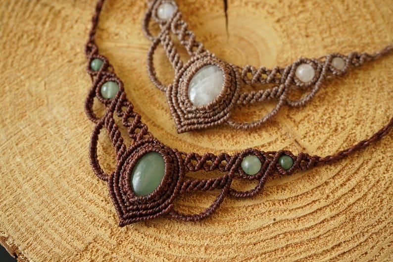 May include: Two handmade macrame necklaces with gemstone accents. One necklace features a large oval green stone, while the other has a large oval white stone. Both are on a wooden surface.