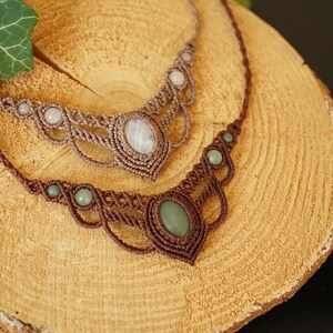 May include: Two macrame necklaces with gemstone accents. One necklace is brown with green stones, the other is tan with white stones. The necklaces are displayed on a wooden surface.