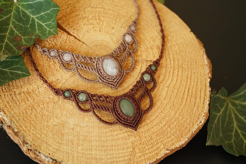 May include: Two macrame necklaces with gemstone accents. One necklace is brown with green stones, the other is tan with white stones. The necklaces are displayed on a wooden surface.