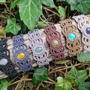 May include: A collection of seven macrame bracelets, each featuring a different gemstone. The bracelets are in various colors, including black, blue, brown, and beige. The gemstones are yellow, green, blue, and purple. The bracelets are displayed on a piece of wood.