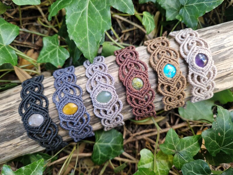 May include: A collection of seven macrame bracelets, each featuring a different gemstone. The bracelets are in various colors, including black, blue, brown, and beige. The gemstones are yellow, green, blue, and purple. The bracelets are displayed on a piece of wood.