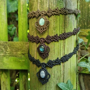 May include: Three macrame necklaces with gemstone pendants are displayed on a weathered wooden post. The necklaces are brown and black, with oval-shaped gemstones in shades of green and blue. The intricate macrame work adds a bohemian touch.