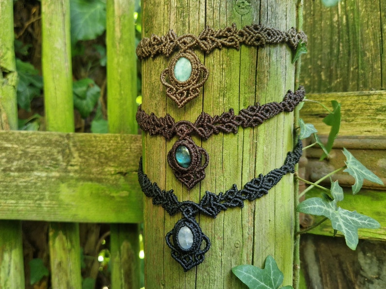 May include: Three macrame necklaces with gemstone pendants are displayed on a weathered wooden post. The necklaces are brown and black, with oval-shaped gemstones in shades of green and blue. The intricate macrame work adds a bohemian touch.