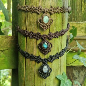May include: Three macrame chokers with gemstone pendants are displayed on a weathered wooden post. The chokers are in shades of brown and black, with intricate knotting patterns. Each features a different gemstone: a light blue, a blue-green, and a white stone.