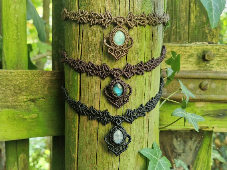 May include: Three macrame chokers with gemstone pendants are displayed on a weathered wooden post. The chokers are in shades of brown and black, with intricate knotting patterns. Each features a different gemstone: a light blue, a blue-green, and a white stone.