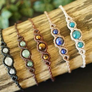 May include: A collection of handmade macrame bracelets featuring various gemstones. The bracelets are displayed on a piece of wood. Gemstones include moonstone, jade, tiger's eye, lapis lazuli, and turquoise. The macrame cord colors vary from black to beige.