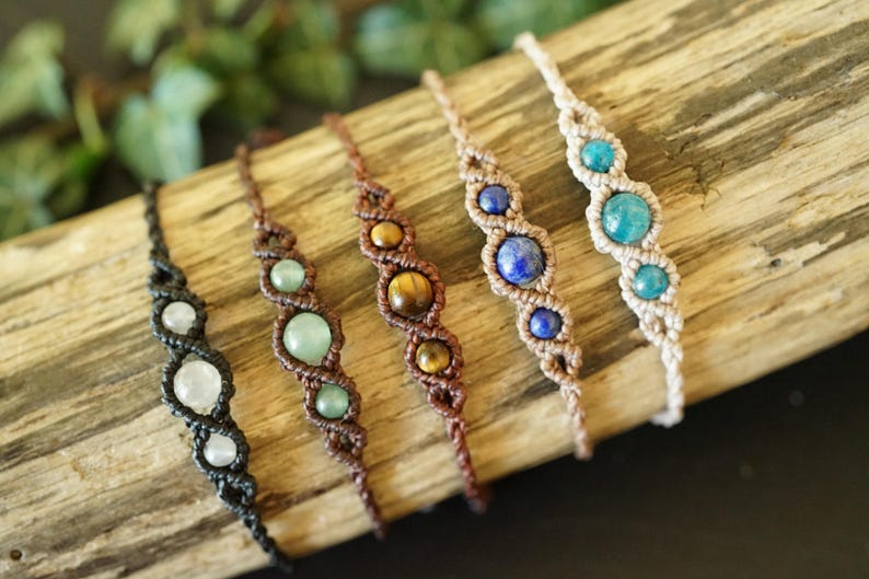 May include: A collection of handmade macrame bracelets featuring various gemstones. The bracelets are displayed on a piece of wood. Gemstones include moonstone, jade, tiger's eye, lapis lazuli, and turquoise. The macrame cord colors vary from black to beige.
