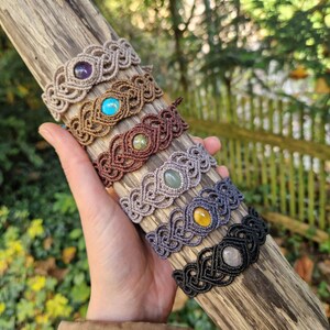 May include: A collection of macrame bracelets in various colors, each featuring a central gemstone. The bracelets are displayed on a piece of driftwood. Gemstones include amethyst, turquoise, and yellow quartz. The macrame is in shades of brown, gray, and black.