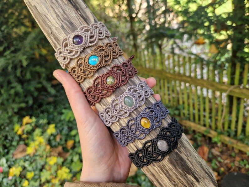 May include: A collection of macrame bracelets in various colors, each featuring a central gemstone. The bracelets are displayed on a piece of driftwood. Gemstones include amethyst, turquoise, and yellow quartz. The macrame is in shades of brown, gray, and black.