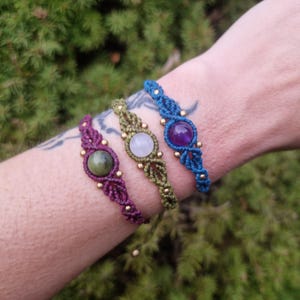 May include: Three macrame bracelets in purple, green, and blue, each featuring a central gemstone and gold-colored beads. The bracelets are displayed on a wrist against a backdrop of green foliage.