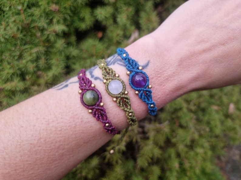 May include: Three macrame bracelets in purple, green, and blue, each featuring a central gemstone and gold-colored beads. The bracelets are displayed on a wrist against a backdrop of green foliage.