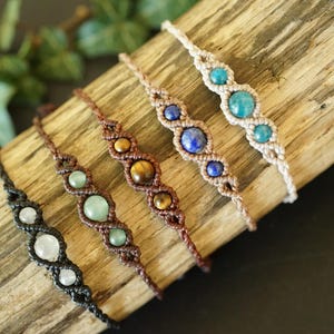 May include: Five handmade macrame bracelets with various gemstone beads, including white, green, tiger's eye, blue, and turquoise. The bracelets are displayed on a piece of wood, showcasing their intricate designs and natural colors.