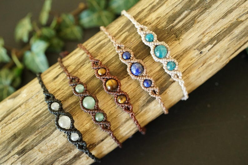 May include: Five handmade macrame bracelets with various gemstone beads, including white, green, tiger's eye, blue, and turquoise. The bracelets are displayed on a piece of wood, showcasing their intricate designs and natural colors.
