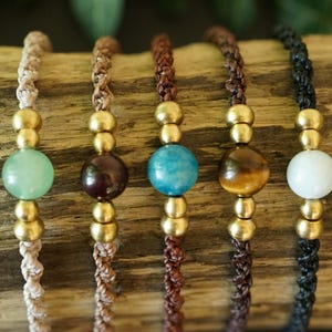 May include: Five handmade bracelets with different colored beads and gold-colored accents. The beads are green, brown, blue, tiger's eye, and white. The bracelets are strung on braided cords in various colors.