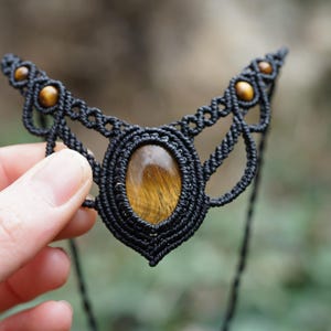 May include: Black macrame necklace with a central oval tiger's eye stone and smaller round stones. The intricate woven design features a symmetrical pattern. The necklace is held against a blurred green background.
