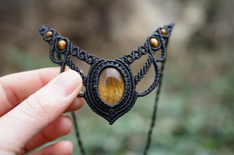 May include: Black macrame necklace with a central oval tiger's eye stone and smaller round stones. The intricate woven design features a symmetrical pattern. The necklace is held against a blurred green background.