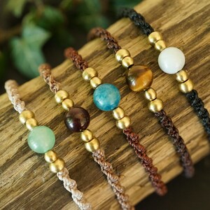 May include: Close-up of five handmade bracelets with various gemstone beads, including green, brown, blue, tiger's eye, and white. Each bracelet features a braided cord and gold-colored metal beads. The bracelets are displayed on a piece of wood.