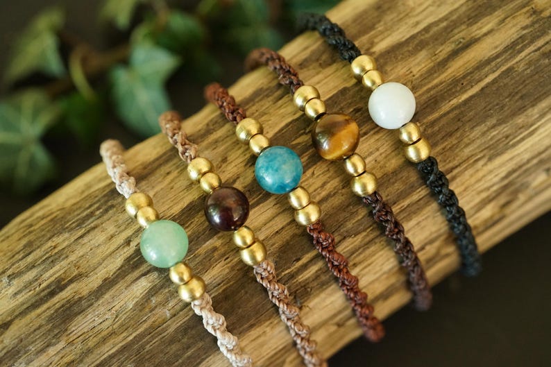 May include: Close-up of five handmade bracelets with various gemstone beads, including green, brown, blue, tiger's eye, and white. Each bracelet features a braided cord and gold-colored metal beads. The bracelets are displayed on a piece of wood.