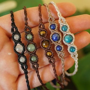 May include: Five macrame bracelets with gemstone beads in various colors, including clear, green, brown, blue, and turquoise. The bracelets are displayed against a neutral background, showcasing the intricate knotting and the natural beauty of the stones.