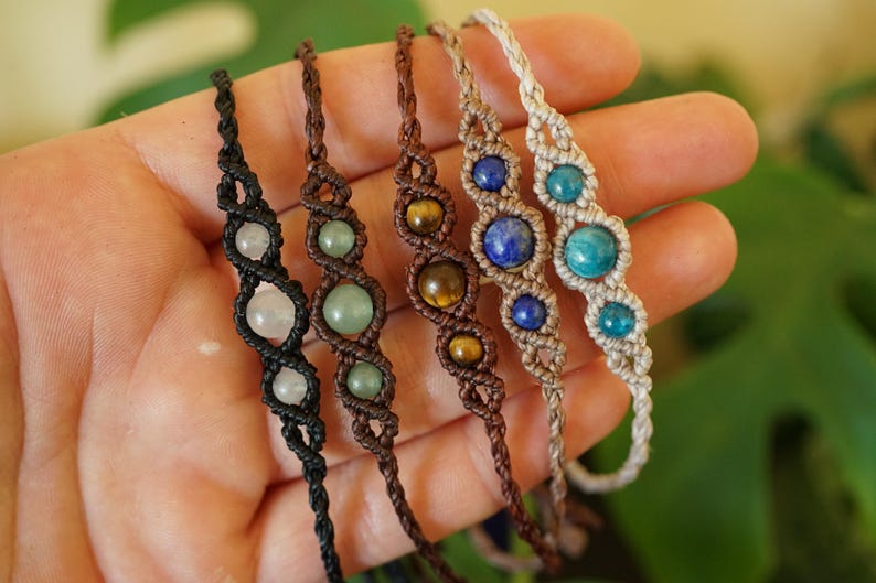 May include: Five macrame bracelets with gemstone beads in various colors, including clear, green, brown, blue, and turquoise. The bracelets are displayed against a neutral background, showcasing the intricate knotting and the natural beauty of the stones.