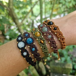 May include: A collection of macrame bracelets in various colors, each featuring different gemstone beads. The bracelets are black, brown, and green, with gold-colored accents. Gemstones include blue, green, and black stones. The bracelets are displayed on a person's arm.