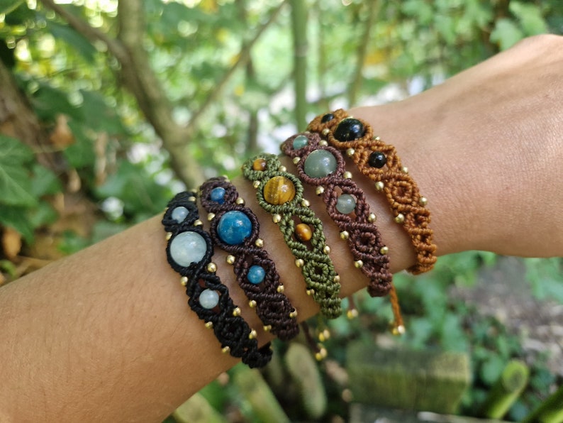 May include: A collection of macrame bracelets in various colors, each featuring different gemstone beads. The bracelets are black, brown, and green, with gold-colored accents. Gemstones include blue, green, and black stones. The bracelets are displayed on a person's arm.
