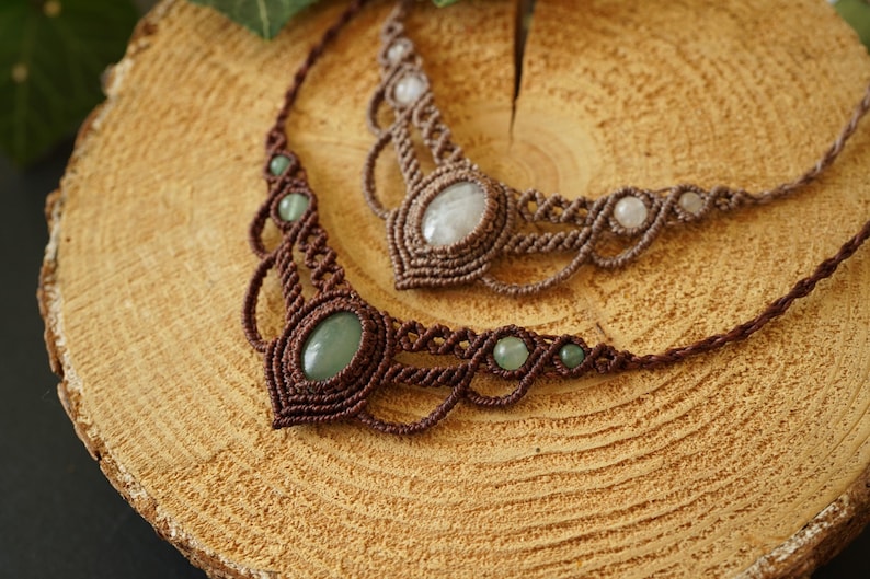 May include: Two handmade macrame necklaces with gemstone accents. One necklace features green stones, the other has white stones. Both are brown and rest on a wooden surface, showcasing the intricate knotting details.