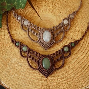 May include: Two macrame necklaces with gemstone pendants. The top necklace is a light brown color with a white oval gemstone. The bottom necklace is a darker brown with a green oval gemstone. Both necklaces are displayed on a wooden surface.