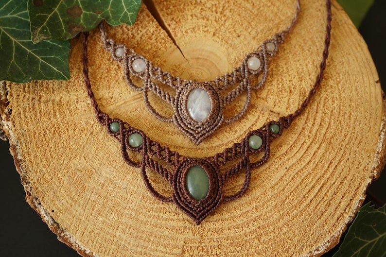 May include: Two macrame necklaces with gemstone pendants. The top necklace is a light brown color with a white oval gemstone. The bottom necklace is a darker brown with a green oval gemstone. Both necklaces are displayed on a wooden surface.