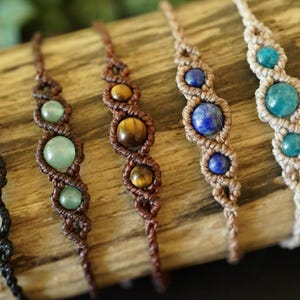 May include: Close-up of six macrame bracelets with gemstone beads. The bracelets feature various colors of gemstones, including white, green, brown, blue, and turquoise. The macrame cord is in shades of black, brown, and beige.