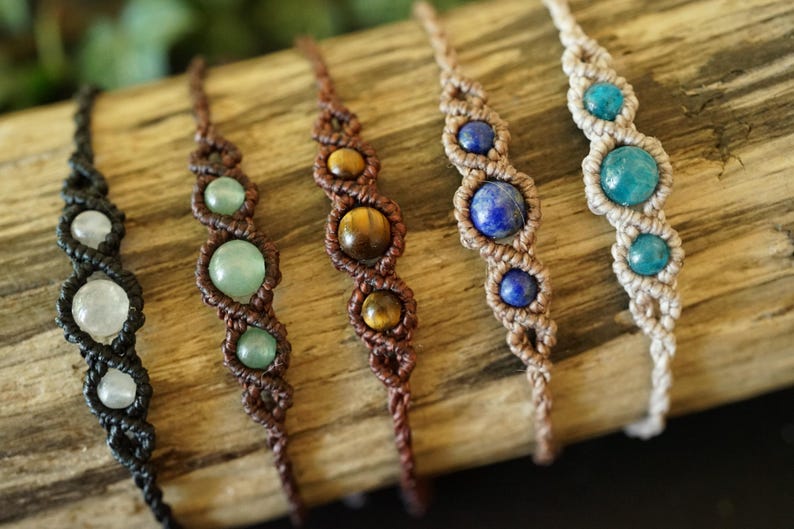 May include: Close-up of six macrame bracelets with gemstone beads. The bracelets feature various colors of gemstones, including white, green, brown, blue, and turquoise. The macrame cord is in shades of black, brown, and beige.