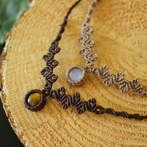 Puede incluir: Dos collares de macram&eacute; con intrincados dise&ntilde;os de cord&oacute;n anudado. Un collar es de color marr&oacute;n oscuro con una piedra preciosa redonda de ojo de tigre, mientras que el otro es de color tostado claro con una piedra redonda, transl&uacute;cida y azul p&aacute;lido.