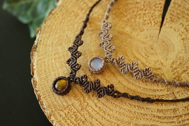 Puede incluir: Dos collares de macram&eacute; con intrincados dise&ntilde;os de cord&oacute;n anudado. Un collar es de color marr&oacute;n oscuro con una piedra preciosa redonda de ojo de tigre, mientras que el otro es de color tostado claro con una piedra redonda, transl&uacute;cida y azul p&aacute;lido.