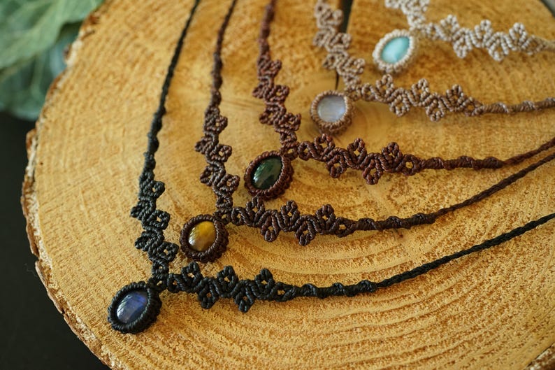 Puede incluir: Cuatro collares de macram&eacute; con dise&ntilde;os de cord&oacute;n anudado en negro, marr&oacute;n oscuro y beige. Cada collar presenta una piedra preciosa ovalada central, incluyendo piedras azules, ojo de tigre, verde oscuro y azul claro, en forma de V.