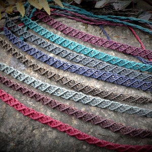Puede incluir: Una colección de collares de macramé hechos a mano en varios colores, incluyendo turquesa, morado y burdeos. Los collares están dispuestos sobre una superficie de piedra, mostrando su intrincado patrón de diamantes y diseño texturizado. La estética general es terrosa y bohemia.