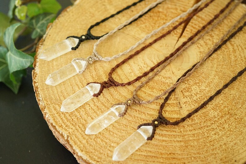 May include: Five necklaces featuring clear, pointed crystal pendants suspended on braided cords in varying shades of black, tan, and brown. Each pendant is secured with a knotted cord design, showcasing the natural transparency of the crystals.
