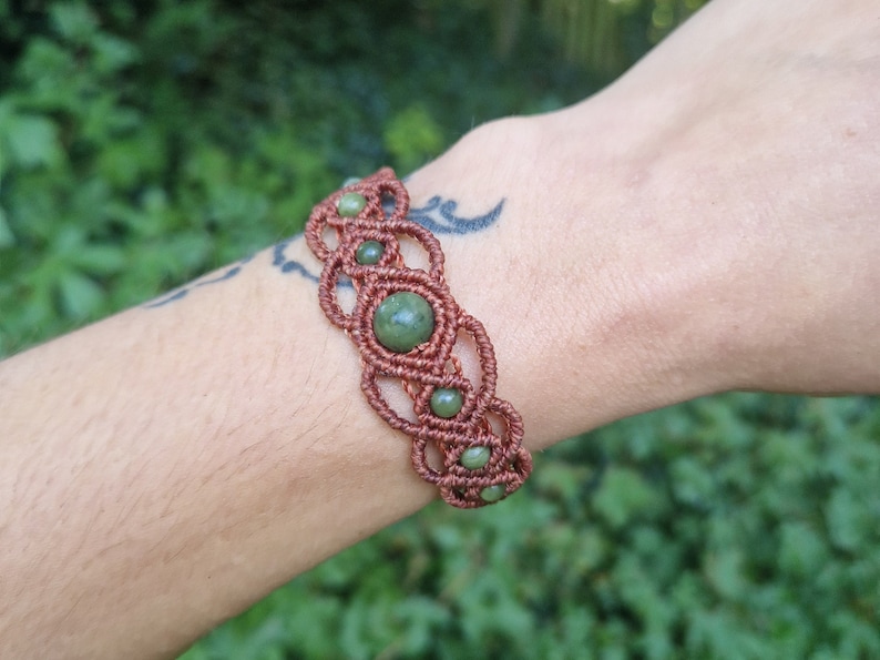 Puede incluir: Pulsera de macram&eacute; hecha a mano en color marr&oacute;n rojizo, con un dise&ntilde;o tejido y cuentas de piedras preciosas verdes. La pulsera se lleva en la mu&ntilde;eca. Las piedras preciosas miden unos 0,8 cm de di&aacute;metro.