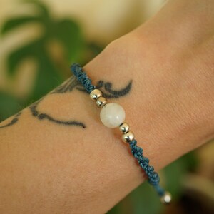 May include: A blue macrame bracelet with a central white gemstone and silver beads. The bracelet is worn on a wrist with a tattoo. The macrame cord is a dark teal color, and the gemstone is round.