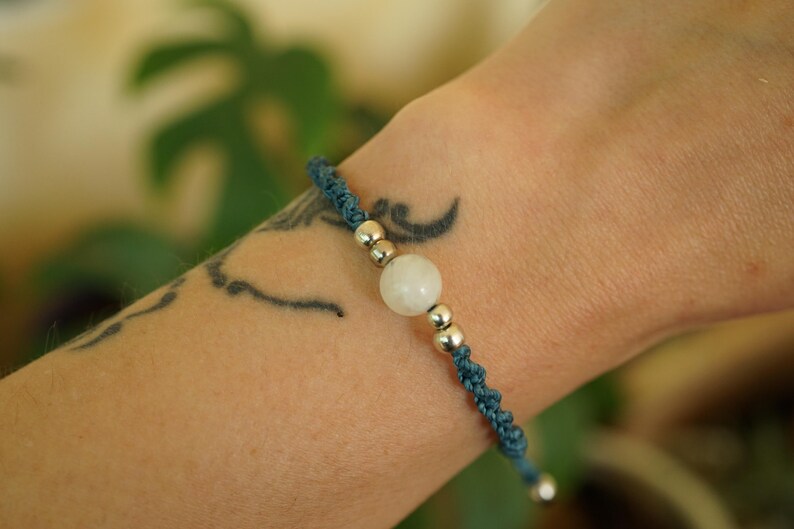 May include: A blue macrame bracelet with a central white gemstone and silver beads. The bracelet is worn on a wrist with a tattoo. The macrame cord is a dark teal color, and the gemstone is round.