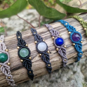 May include: A collection of macrame bracelets with various gemstone centerpieces. The bracelets feature a woven design in colors like black, gray, blue, green, and purple. Each bracelet has a different gemstone, including malachite, amethyst, and lapis lazuli.