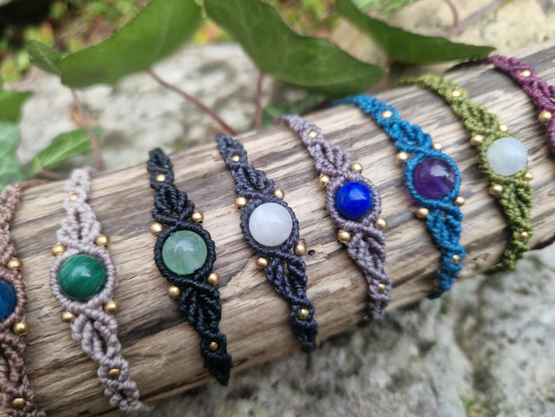 May include: A collection of macrame bracelets with various gemstone centerpieces. The bracelets feature a woven design in colors like black, gray, blue, green, and purple. Each bracelet has a different gemstone, including malachite, amethyst, and lapis lazuli.