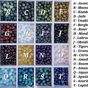 May include: A wooden tray filled with various gemstone beads, each in a separate compartment. The beads are in a variety of colors, including green, brown, white, black, yellow, purple, blue, and pink. Each compartment is labeled with a letter and a gemstone name.