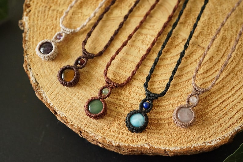 May include: Close-up of several macrame necklaces with gemstone pendants. The necklaces feature various colors of cord and gemstones, including amethyst, citrine, green aventurine, blue lapis lazuli, and rose quartz. The necklaces are displayed on a wooden surface.