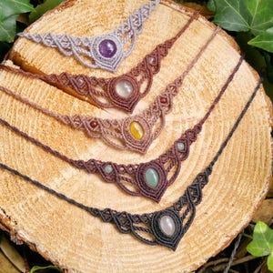 Op de afbeelding: Vijf macramé kettingen met edelsteen hangers worden getoond op een houten oppervlak. De kettingen hebben verschillende kleuren koord en edelstenen, waaronder amethist, kwarts en jade. De kettingen zijn in een rij gerangschikt.