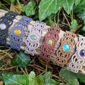 May include: A collection of seven macrame bracelets in various colors, each featuring a central gemstone. The bracelets are displayed on a piece of wood, surrounded by green foliage. Gemstones include a white, yellow, green, brown, turquoise, and purple stone.
