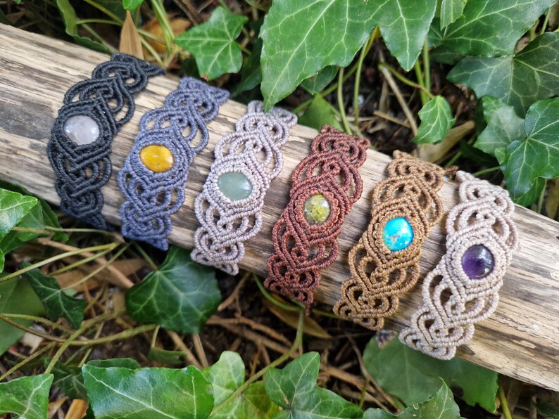 May include: A collection of seven macrame bracelets in various colors, each featuring a central gemstone. The bracelets are displayed on a piece of wood, surrounded by green foliage. Gemstones include a white, yellow, green, brown, turquoise, and purple stone.